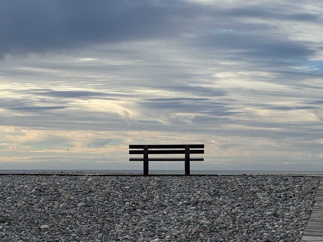 Eine leere Bank auf einem Kieselstrand, dahinter das Meer und ein weiter Wolkenhimmel