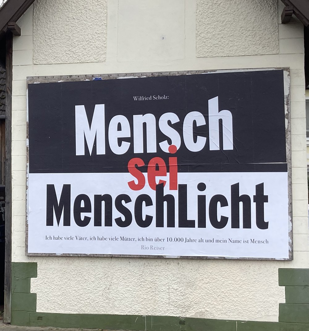 Ein Plakat an einer Hauswand: „Mensch sei MenschLicht“, darunter ein Zitat von Rio Reiser — „Ich habe viele Väter, ich habe viele Mütter, ich bin über 10.000 Jahre alt und mein Name ist Mensch“
